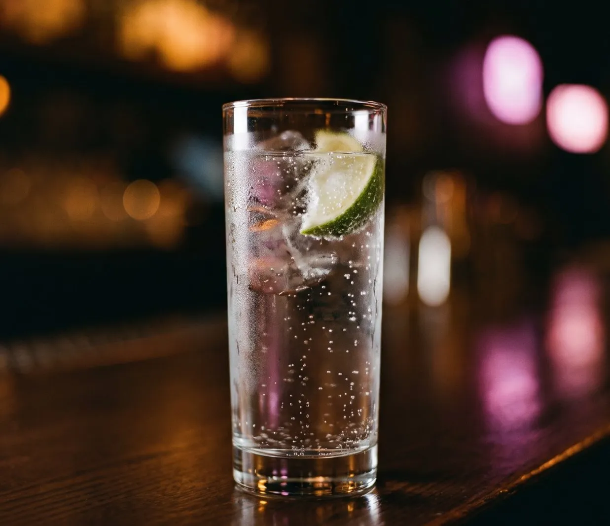 Hohes Glas Sprudelwasser mit Eis und Limettenspalte auf einem dunklen Holztresen, dahinter unscharfe bernsteinfarbene und rosa Barlichter