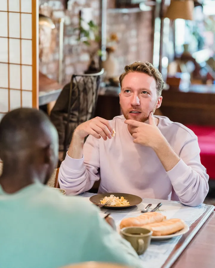 Zwei Männer beim Frühstück am Holztisch, einer im fliederfarbenen Pullover blickt nachdenklich zur Seite, der andere sitzt mit dem Rücken zur Kamera, davor Teller und Kaffeetasse