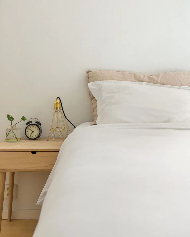Aufgeräumtes Schlafzimmer bei Tageslicht, weiße Bettwäsche vor einem beigen Leinenkissen, daneben ein Holznachttisch mit Drahtlampe, Wecker und einem Steckling im Glas