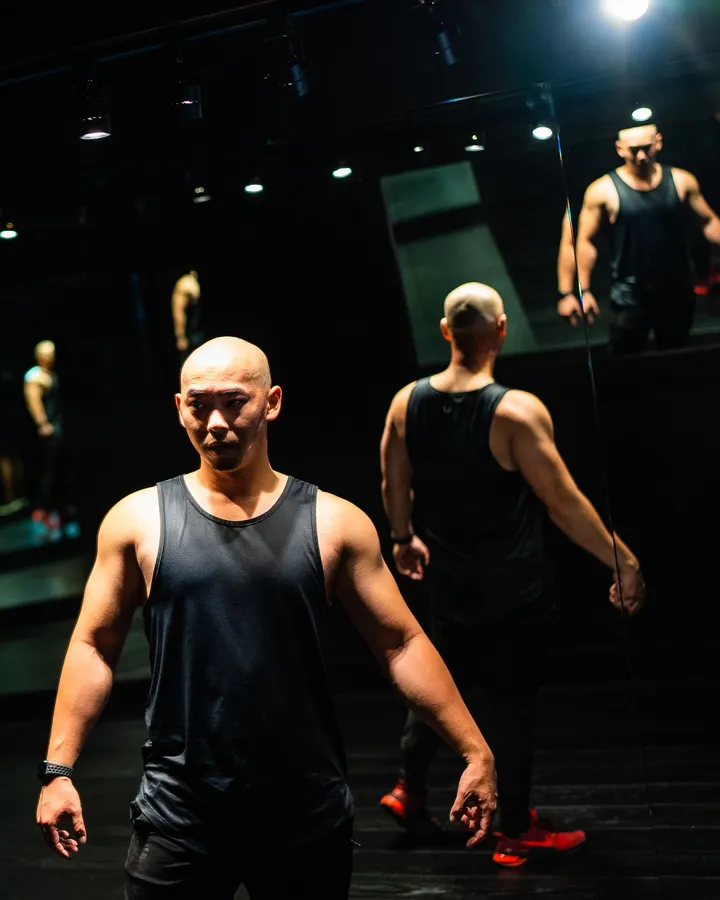 Muskulöser Mann im schwarzen Tanktop in einem dunklen Trainingsraum, hinter ihm wirft eine Spiegelwand sein Bild mehrfach zurück